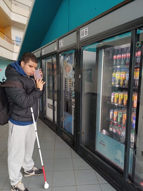 Un jeune homme malvoyant se tient face à des distributeurs automatiques de boissons avec son téléphone